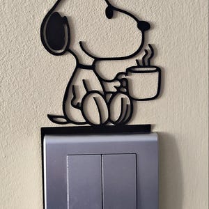 Könnte beinhalten: Ein schwarzer, flacher Wandaufkleber in Silhouettenform, der einen sitzenden Cartoon-Hund mit einer dampfenden Tasse zeigt. Der Aufkleber ist direkt auf einer rechteckigen, silberfarbenen Doppel-Lichtschalterabdeckung an der Wand angebracht.