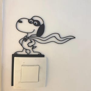 May include: A black metal Snoopy-inspired light switch cover. The design features Snoopy in a superhero pose with a flowing cape. The cover is mounted above a white light switch on a white wall.