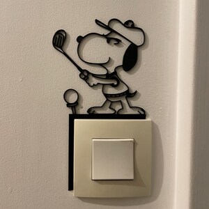 Op de afbeelding: Zwarte metalen Snoopy-figuur in een golfhouding, gemonteerd boven een lichtschakelaar. De figuur is een silhouet, met Snoopy die een golfhoed draagt en een golfclub vasthoudt. Er is ook een golfbal aanwezig.