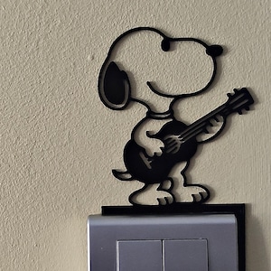 Può includere: Una sagoma nera piatta o ritaglio del personaggio dei cartoni animati Snoopy che suona la chitarra. La figura è posizionata in piedi sopra una placca rettangolare per interruttore della luce di colore argento montata su una parete beige.