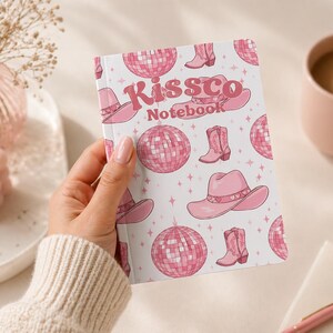 Op de afbeelding: Een notitieboek met een roze en wit patroon met cowboylaarzen, discoballen en cowboyhoeden. Het notitieboek heeft de tekst "Kissco Notebook" op de kaft staan. Het notitieboek wordt in de hand van een persoon gehouden.