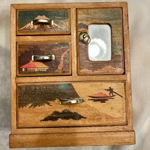 May include: A wooden chest with three drawers and one cabinet door. The drawer fronts feature inlaid wood scenes of Mount Fuji, a building, and a boat with a person. The cabinet door has a small rectangular mirror and a circular metal pull.