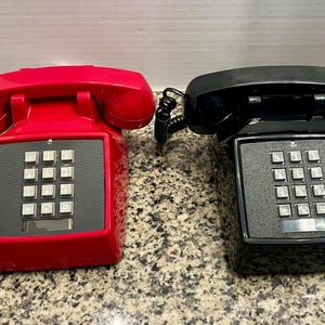 May include: Two desk telephones, one in bright red and one in glossy black, each featuring a matching handset, a coiled cord, and a square keypad with twelve silver-colored buttons set against a dark gray panel on the front of the base.
