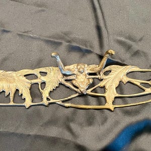 May include: A vintage brass wall-mounted coat rack. The rack features four hooks with an ornate design of leaves and figures. The brass has a warm, aged patina. The overall length is approximately 24 inches.