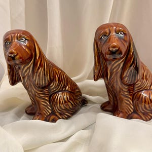 May include: Two glossy ceramic figurines of brown spaniel dogs in a sitting position. Each dog features long, textured ears, dark brown fur detailing, and painted eyes with a downward gaze, rendered in a shiny, reflective glazed finish.