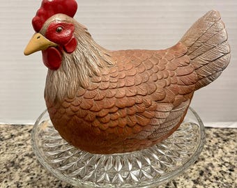 May include: A brown ceramic chicken figurine with a red comb and wattle and a yellow beak, resting on a clear, circular glass pedestal dish with a diamond-patterned base. The chicken features detailed, textured feather carvings across its body.