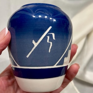 May include: A small ceramic vase featuring a deep blue upper section that transitions into a lighter blue tone. The surface is decorated with white hand-painted line art and geometric patterns, finished with a solid white base and a dark blue band.