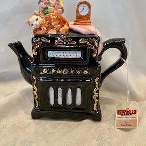 May include: A black ceramic teapot shaped like a vintage radio with gold accents. The lid features a sleeping orange cat, a small clock, and a bouquet of flowers. A tag attached to the handle reads TEA NEE TEA POT Handpainted Designed Collectible by Cardinal.