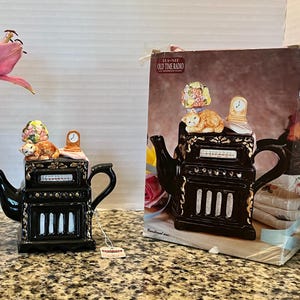 May include: A black ceramic teapot shaped like an old-time radio with gold accents. The lid features a sleeping orange cat, a small clock, and a floral bouquet. The teapot is accompanied by its original cardboard box labeled Tea-Nee Old Time Radio.