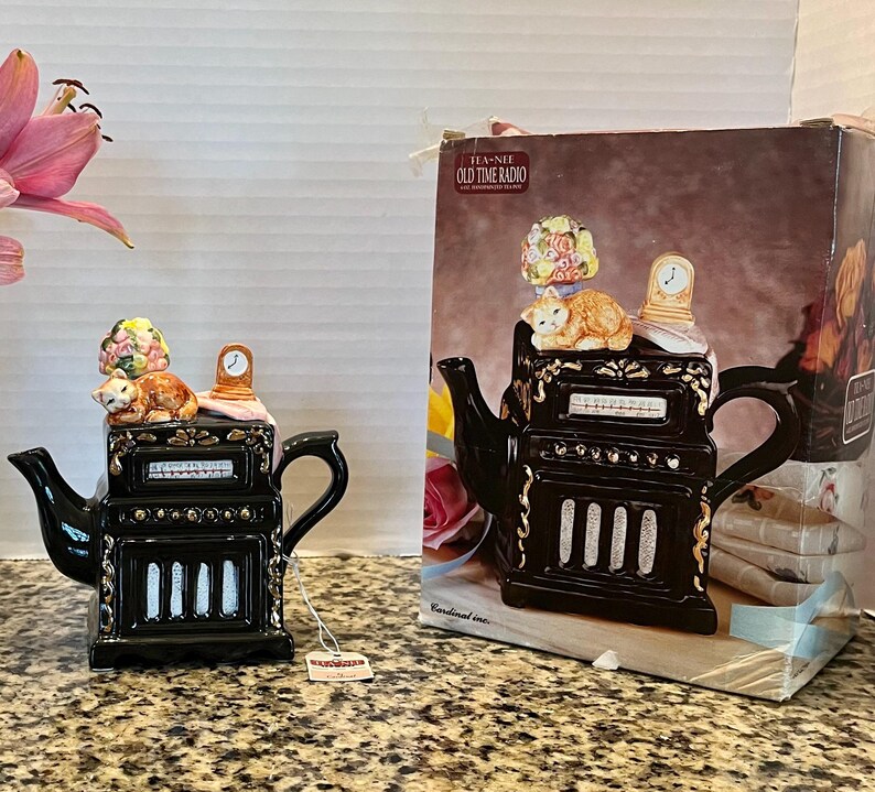 May include: A black ceramic teapot shaped like an old-time radio with gold accents. The lid features a sleeping orange cat, a small clock, and a floral bouquet. The teapot is accompanied by its original cardboard box labeled Tea-Nee Old Time Radio.