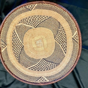May include: A round woven basket with a brown rim and a spiral design. The basket features alternating patterns of tan and black woven fibers, creating a textured surface. The center is a lighter tan color.