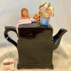 May include: A glossy black ceramic teapot featuring a sculpted lid ornament of a pink hat and a colorful bouquet of flowers. The teapot has a curved handle and spout, and a white tag attached by a string reads TEA NEE TEA POT by Cardinal.