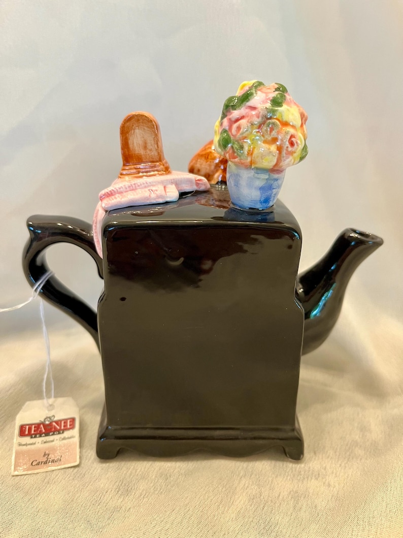May include: A glossy black ceramic teapot featuring a sculpted lid ornament of a pink hat and a colorful bouquet of flowers. The teapot has a curved handle and spout, and a white tag attached by a string reads TEA NEE TEA POT by Cardinal.