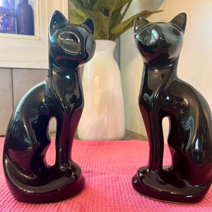 May include: Two glossy black ceramic cat figurines. The cats are in a seated position with their tails curled around their bodies. The figurines have a sleek, modern design and are displayed on a pink cloth. A vase of sunflowers and other decorative items are in the background.