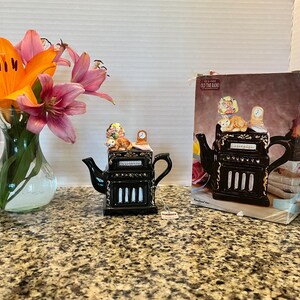 May include: A black ceramic teapot shaped like an old-time radio with gold accents, featuring a cat and a lamp on top. The teapot is displayed next to its original cardboard box, which shows the same radio design and the text TEA-NEE OLD TIME RADIO.