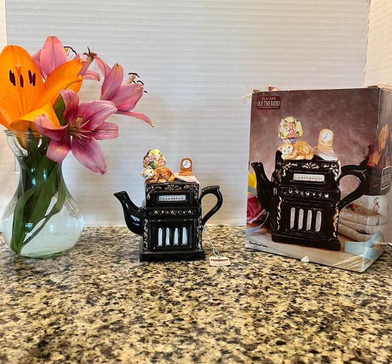 May include: A black ceramic teapot shaped like an old-time radio with gold accents, featuring a cat and a lamp on top. The teapot is displayed next to its original cardboard box, which shows the same radio design and the text TEA-NEE OLD TIME RADIO.