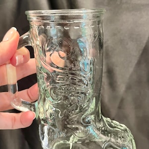 May include: A clear glass mug shaped like a cowboy boot, featuring an embossed decorative pattern across the surface. The mug includes a sturdy handle attached to the side, with a wide rim at the top and a detailed molded design resembling stitching.