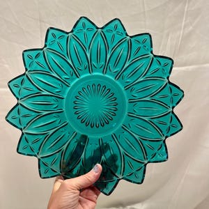 May include: A circular teal glass plate featuring a sunburst pattern with radiating petals and a central starburst design. The edges of the plate are scalloped, creating a jagged, star-like silhouette with a translucent finish and pressed glass texture.