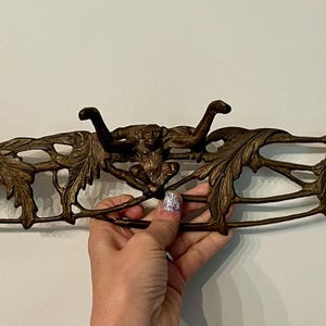 May include: An antique bronze wall-mounted coat rack with three hooks. The rack features an ornate design with leaves and bull heads. The hooks are curved and the overall design is symmetrical. The rack is held by a hand.