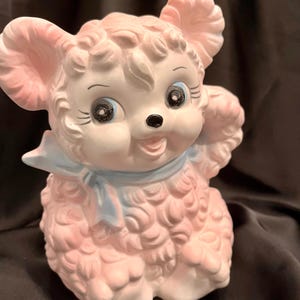 May include: A ceramic figurine of a lamb with curly pink fur, large blue eyes, and long eyelashes. The lamb has a light blue ribbon tied around its neck and a small black nose. The figure is sculpted with a textured, woolly appearance.