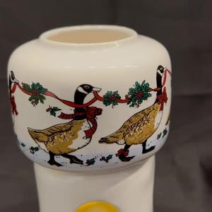 May include: A cream-colored ceramic oil warmer featuring a cylindrical base with a circular opening and a wider top bowl. The bowl is decorated with a repeating pattern of geese wearing red bows, connected by green holly garlands and red ribbons.