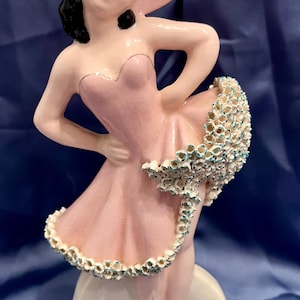 May include: A ceramic figurine of a person wearing a pink dress and a matching wide-brimmed hat. The figure has black hair, red lips, and black shoes. The dress features a textured, ruffled hemline with small white and teal floral-like details.