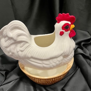 May include: A white ceramic chicken-shaped container with a red comb and wattle, a yellow beak, and black eyes. The chicken rests on a separate, tan-colored ceramic base designed with a textured, woven basket-weave pattern on the exterior.