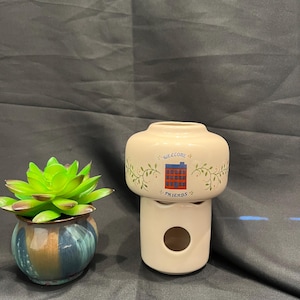 May include: A cream-colored ceramic oil warmer featuring a graphic of a red building with blue windows and the text "WELCOME FRIENDS" surrounded by green vines, positioned next to a small, round, multi-colored succulent planter with blue and brown tones.