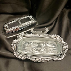 May include: A silver-plated butter dish featuring a rectangular glass insert with a sunburst pattern at the base. The metal frame has an ornate, embossed border with shell-like motifs and a matching silver lid with a decorative handle.