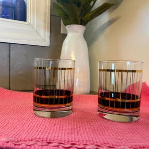 May include: Two vintage drinking glasses with a black and gold striped design. The glasses have a thick, dark base and a gold rim. They are sitting on a pink cloth.