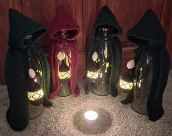 May include: Four clear glass bottles are each draped in a hand-knitted hooded cape, three in dark green and one in deep red. Inside each bottle, a small, glowing, acorn-shaped light hangs. A single tea light candle sits in the centre.