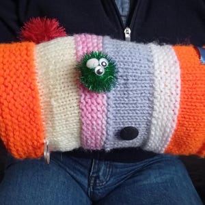 May include: A knitted arm warmer in a striped pattern of orange, cream, pink, and blue. It has a green, fuzzy decoration with googly eyes, a red pom-pom, a black button, and a small silver ring. The design is handmade.