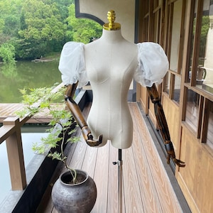 May include: A beige dress form with wooden articulated arms and a gold-colored neck cap. The form is wearing white, puffy sleeves. It stands on a black tripod base on a wooden deck, with a potted plant and a lake in the background.