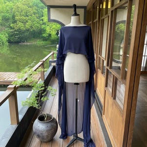 以下が含まれることがあります： マネキンに着せられたネイビーブルーのツーピースのフォーマルな衣装。トップスは、重ね着された袖が付いた、ボートネックのクロップドスタイルです。スカートは長く流れるようなデザインです。衣装は、湖と木々が見える木製のデッキに展示されています。