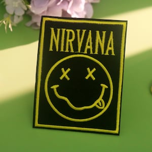 Puede incluir: Parche rectangular negro con la palabra "NIRVANA" en amarillo, sobre una carita sonriente amarilla con los ojos tachados y la lengua fuera. El parche está sobre una superficie verde, con dos piruletas coloridas y flores en el fondo.