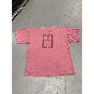 May include: A faded red short-sleeve t-shirt featuring a rectangular graphic on the center chest. The graphic includes the text KK, Krispy Kreme, Original Glazed, and additional small print surrounded by a border of small star icons.