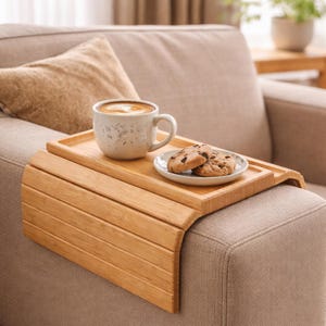 May include: A bamboo sofa arm tray holds a mug of coffee and a small plate with two chocolate chip cookies. The tray is light brown and rests on the arm of a beige sofa. A beige pillow is in the background.
