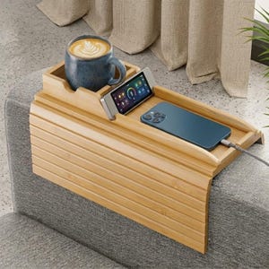 May include: A bamboo sofa arm tray with a built-in cup holder, a small device holder, and a larger area for a phone. A blue mug with a latte sits in the cup holder. A blue phone is plugged in. The tray is on a gray sofa.