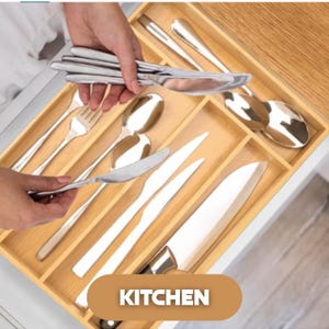 May include: A wooden cutlery organizer filled with stainless steel silverware. The organizer is inside a drawer, with forks, spoons, and knives neatly arranged. The word "KITCHEN" is displayed in a brown rectangle.