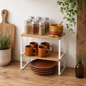 May include: A two-tiered bamboo and white metal kitchen shelf displaying various kitchen items. The top shelf holds glass spice jars, and ceramic bowls. The bottom shelf has brown mugs and plates. A wooden cutting board and plant are in the background.