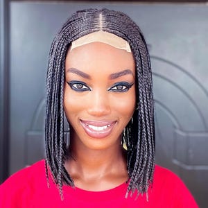 May include: A smiling person with a bob-length braided wig, wearing a red top. The wig features small, tightly woven braids and a natural-looking hairline. The person has dark eyeliner and a bright smile.