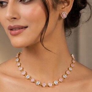 May include: A gold-toned necklace featuring a series of oval-shaped light pink stones surrounded by small clear crystals, paired with matching circular stud earrings that have a central pink stone encircled by a halo of clear crystals.