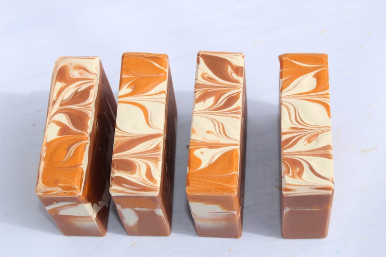 Op de afbeelding: Vier rechthoekige handgemaakte zeepblokken met een wervelend ontwerp. De zeep heeft tinten bruin, oranje en cr&egrave;me. De blokken zijn verticaal geplaatst tegen een witte achtergrond, waarbij hun unieke patronen worden getoond.