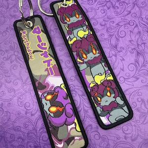 May include: Two rectangular keychains with metal rings. One features the text "Marshadow" and Japanese characters on a camouflage background. The other keychain displays a repeating cartoon character with purple, gray, and yellow accents.