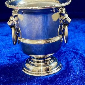 Op de afbeelding: Een zilverkleurige wijnkoeler met een brede rand en een voet. De koeler heeft twee leeuwenkopgrepen met ringen. De glanzende metalen koeler wordt gepresenteerd tegen een blauwe achtergrond. Geschatte hoogte: 15 cm.