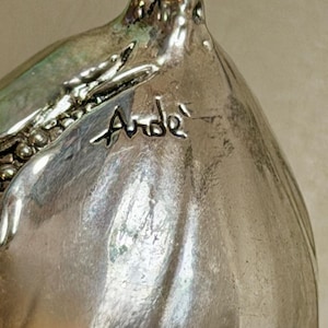 Può includere: Un oggetto metallico color argento lucido con una forma organica curva e un bordo testurizzato a perline. La superficie presenta il testo inciso Arde in corsivo, con sottili riflessi e punti luce sul metallo liscio.