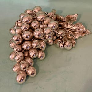 Può includere: Una scultura a grappolo d'uva color argento con foglie dettagliate. L'uva è sferica e riflette la luce, creando un aspetto lucido. La scultura è probabilmente un oggetto decorativo per una casa o una cucina.