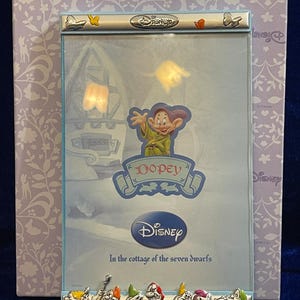Op de afbeelding: Een Disney-fotolijst met het personage Dopey uit Sneeuwwitje. De lijst heeft een zilveren rand met het Disney-logo en de tekst "In the cottage of the seven dwarfs". De achtergrond is lichtpaars met een bloemenpatroon.
