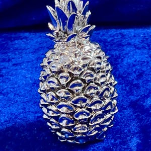 Może przedstawiać: Srebrna rzeźba ananasa o szczegółowej fakturze. Ananas ma teksturowany korpus i kolczastą koronę. Rzeźba jest umieszczona na głębokim niebieskim tle, tworząc uderzający kontrast.