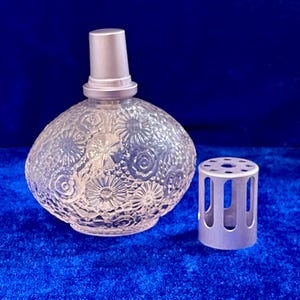 May include: A clear glass perfume bottle with a floral pattern and a silver-colored cap. The bottle is round and has a matching silver-colored decorative sleeve. The bottle is sitting on a blue surface.